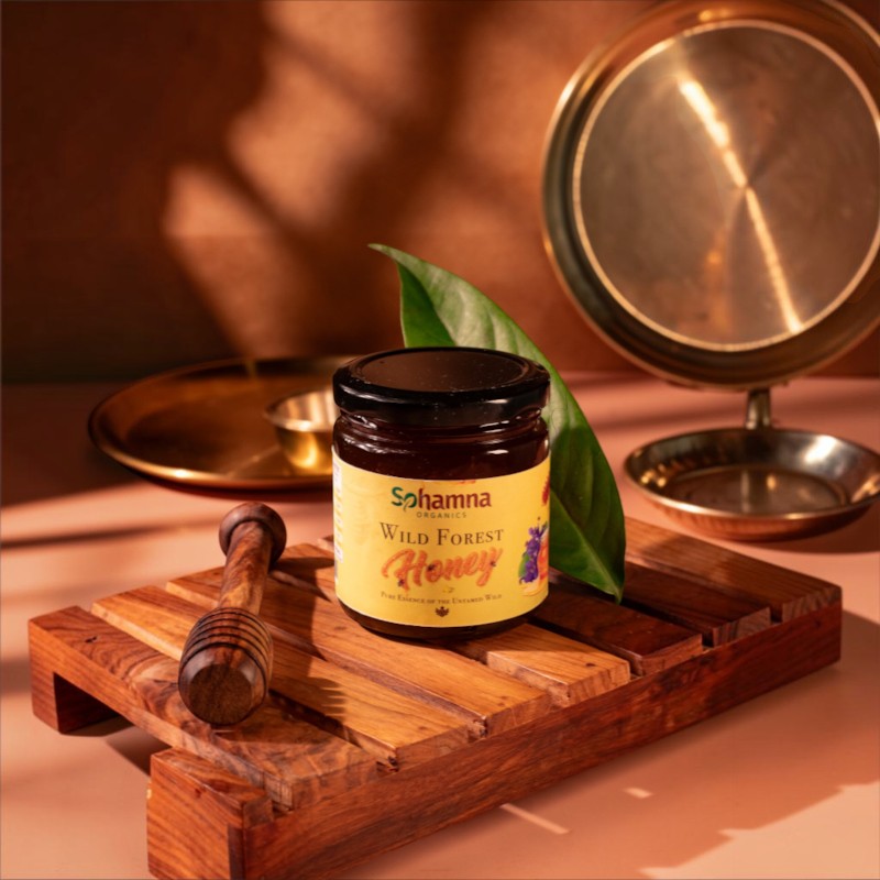 A jar of Sohamna Wild Forest Honey sits on a wooden tray with a wooden honey dipper and a green leaf, set against a warm, rustic background with brass dishes.