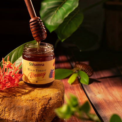 A jar of Sohamna Organics Wild Forest Honey sits on a rustic stone block with a wooden honey dipper dripping honey into it. The scene is styled with green leaves and orange flowers on a dark wooden table.
