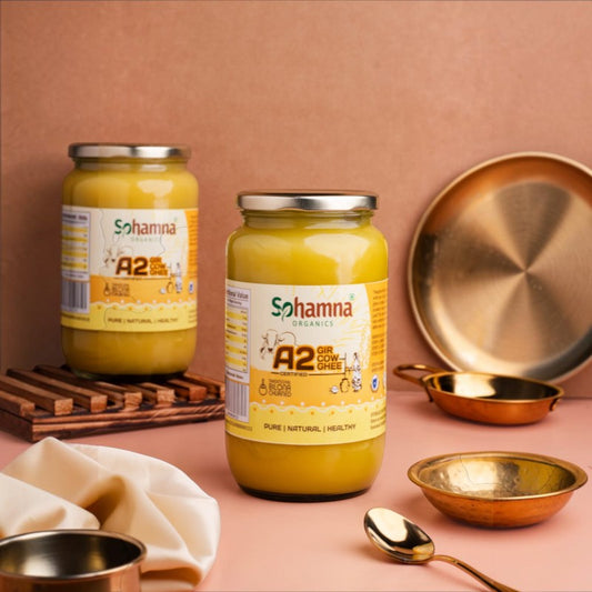 Two glass jars of Sohamna Organics A2 Curd Greese are displayed on a pink surface with a matching pink background, surrounded by decorative brass and copper bowls, a spoon, and a wooden tray.