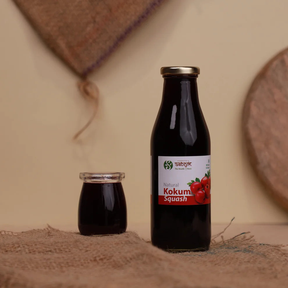 A bottle of Natural Kokum Squash next to a small glass jar, both filled with dark brown liquid, set on a rustic burlap surface against a warm beige background.