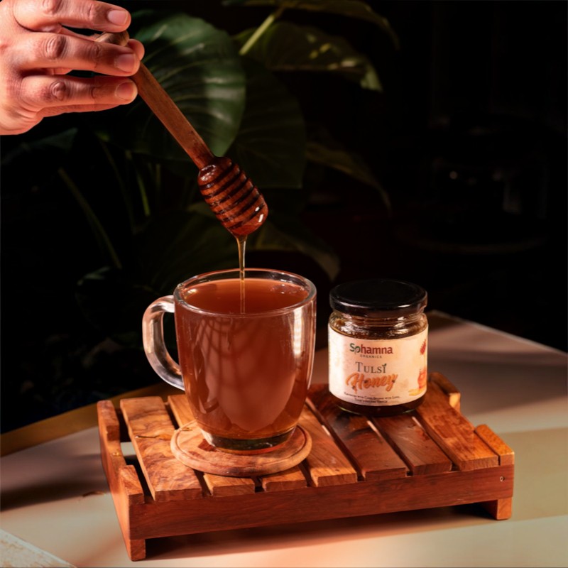 A hand holds a wooden honey dipper above a clear glass mug filled with amber honey, with a jar of Sohamna Tulsi Honey beside it on a wooden tray against a dark leafy background.