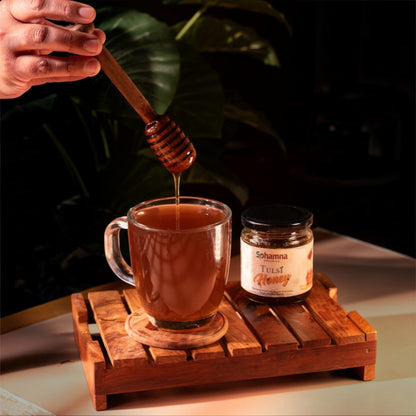 A hand holds a wooden honey dipper above a clear glass mug filled with amber honey, with a jar of Sohamna Tulsi Honey beside it on a wooden tray against a dark leafy background.