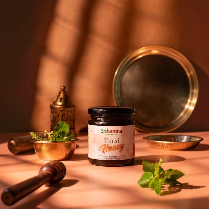 A jar of Sohamna Tulsi Honey is displayed on a warm, peach-colored surface with a rustic background. The scene is styled with traditional Indian elements, including brass dishes, a wooden honey dipper, and fresh green tulsi leaves, all illuminated by warm, directional sunlight casting soft shadows.