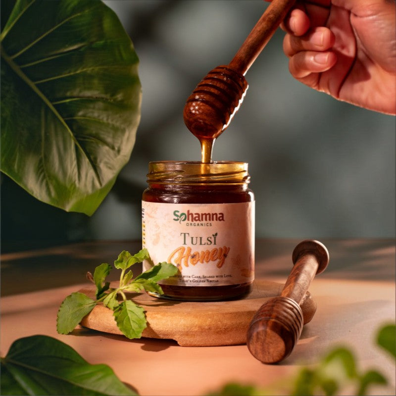 A jar of Sohamna Organics Tulsi Honey sits on a wooden board with honey dripping from a wooden dipper held by a hand. Fresh green leaves and a second dipper are arranged around it on a warm, softly lit surface.