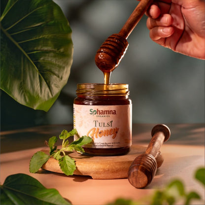 A jar of Sohamna Organics Tulsi Honey sits on a wooden board with honey dripping from a wooden dipper held by a hand. Fresh green leaves and a second dipper are arranged around it on a warm, softly lit surface.
